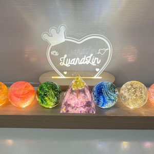 Pode incluir: Um letreiro luminoso em forma de coração com o nome "LyandLin" e uma coroa no topo, rodeado por sete orbes de vidro coloridos em uma base de madeira.
