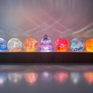 Pode incluir: Sete orbes de vidro com padrões de cores em espiral, iluminados por dentro, sentam-se sobre uma base de madeira. Uma pirâmide de vidro transparente com uma flor roxa dentro se senta no centro.
