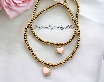 May include: Two gold beaded bracelets with pink heart charms. The bracelets are layered on top of each other. The text "Bijoux Bijoux by Alex" is visible in the image.