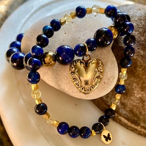 Può includere: Due braccialetti con perline blu e dorate. Un braccialetto ha un ciondolo a forma di cuore dorato e l'altro ha un ciondolo a forma di farfalla dorato.