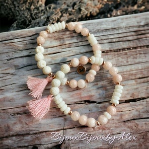 Pode incluir: Duas pulseiras com contas rosa e brancas, uma com um amuleto de borboleta e a outra com borlas rosa. As pulseiras estão sobre uma superfície de madeira.