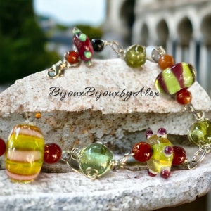 May include: A silver chain bracelet with colourful glass beads. The beads are green, yellow, red and white. The bracelet is on a rock with the text "Bijoux Bijoux by Alex" on it.