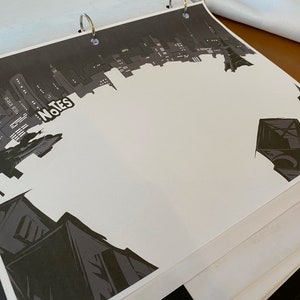 May include: A black and white illustration of a city skyline with the word "Notes" written in white on a black background. The image is printed on a white sheet of paper in a binder.