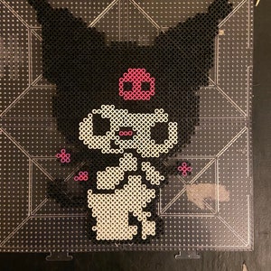 May include: A pixel art design of a cartoon character with black fur, large ears, and a pink skull-shaped emblem. The character is made of black, white, and pink beads on a clear pegboard, creating a unique, handmade aesthetic.