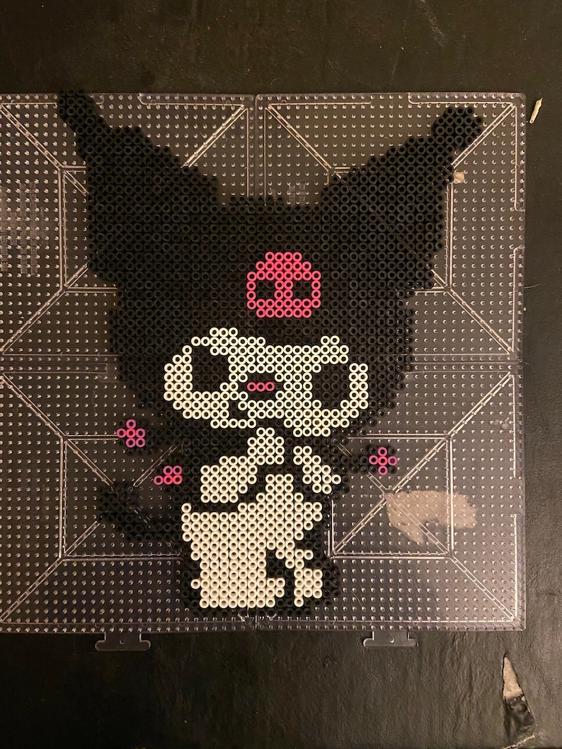 May include: A pixel art design of a cartoon character with black fur, large ears, and a pink skull-shaped emblem. The character is made of black, white, and pink beads on a clear pegboard, creating a unique, handmade aesthetic.