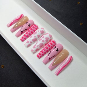 May include: Set of pink and white press-on nails with different designs, including cow print, polka dots, and a smiley face with an X for eyes.