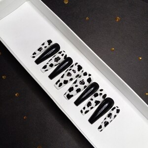 May include: A set of ten black and white cow print press-on nails in a white box. The nails are long and have a glossy finish.