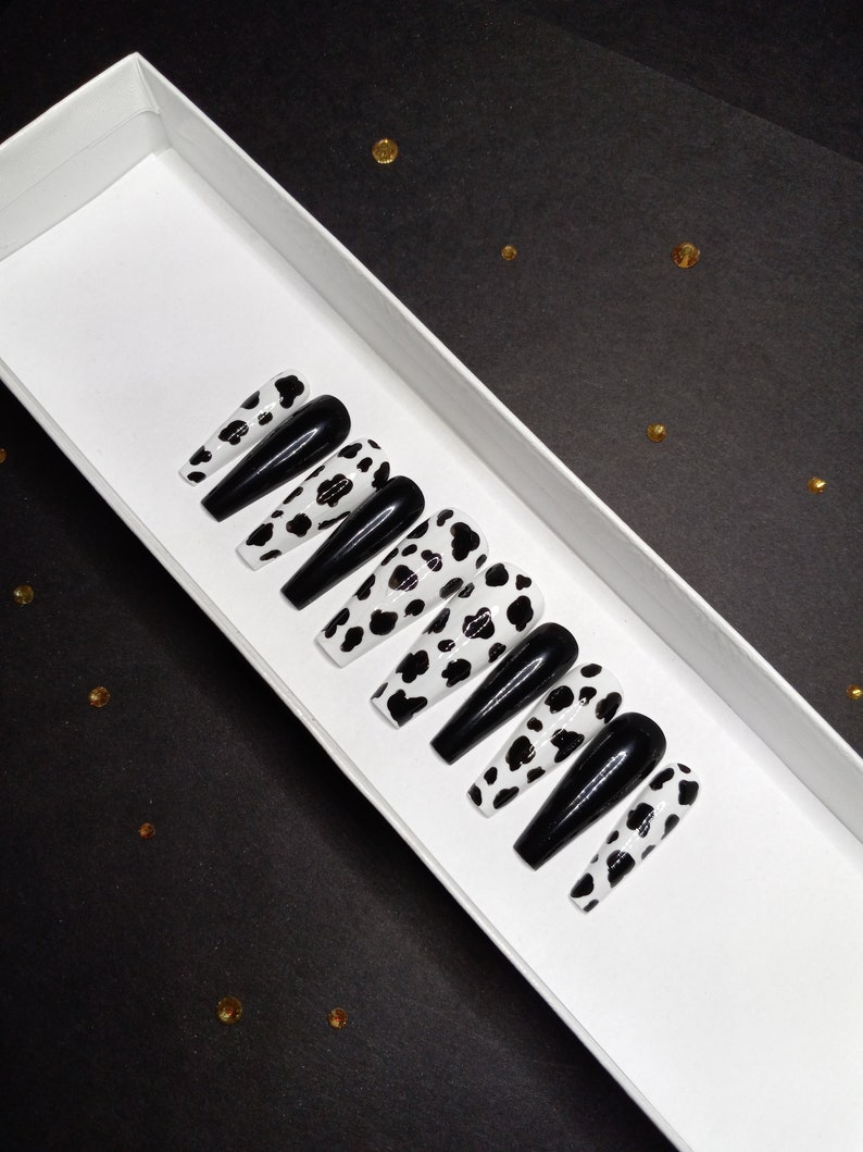 May include: A set of ten black and white cow print press-on nails in a white box. The nails are long and have a glossy finish.