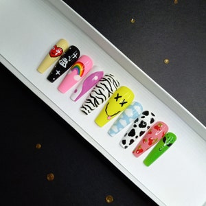 May include: A set of ten coffin-shaped press-on nails featuring diverse designs including a red heart on yellow, a black alien face, a rainbow, purple swirls, black and white zebra stripes, a yellow smiley face, blue clouds, cow print, cherries, and green.