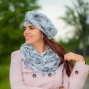 May include: A woman wearing a pink coat with a gray and white crocheted beret and scarf. The scarf has small pom-poms around the edges.