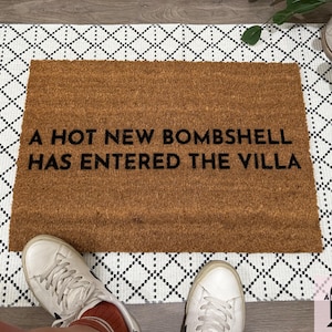 May include: A rectangular doormat made of brown coir fiber, with the phrase "A HOT NEW BOMBSHELL HAS ENTERED THE VILLA" in black lettering. The mat is placed on a white rug with a black diamond pattern. White sneakers are visible at the bottom.