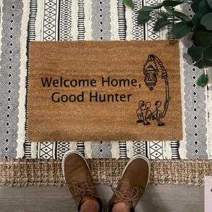 May include: A brown coir doormat with the text "Welcome Home, Good Hunter" and a black and white illustration of a lantern and three figures.
