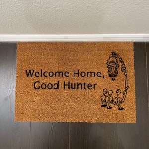 May include: A brown coir doormat with the text "Welcome Home, Good Hunter" and a black silhouette of a lantern and three figures.