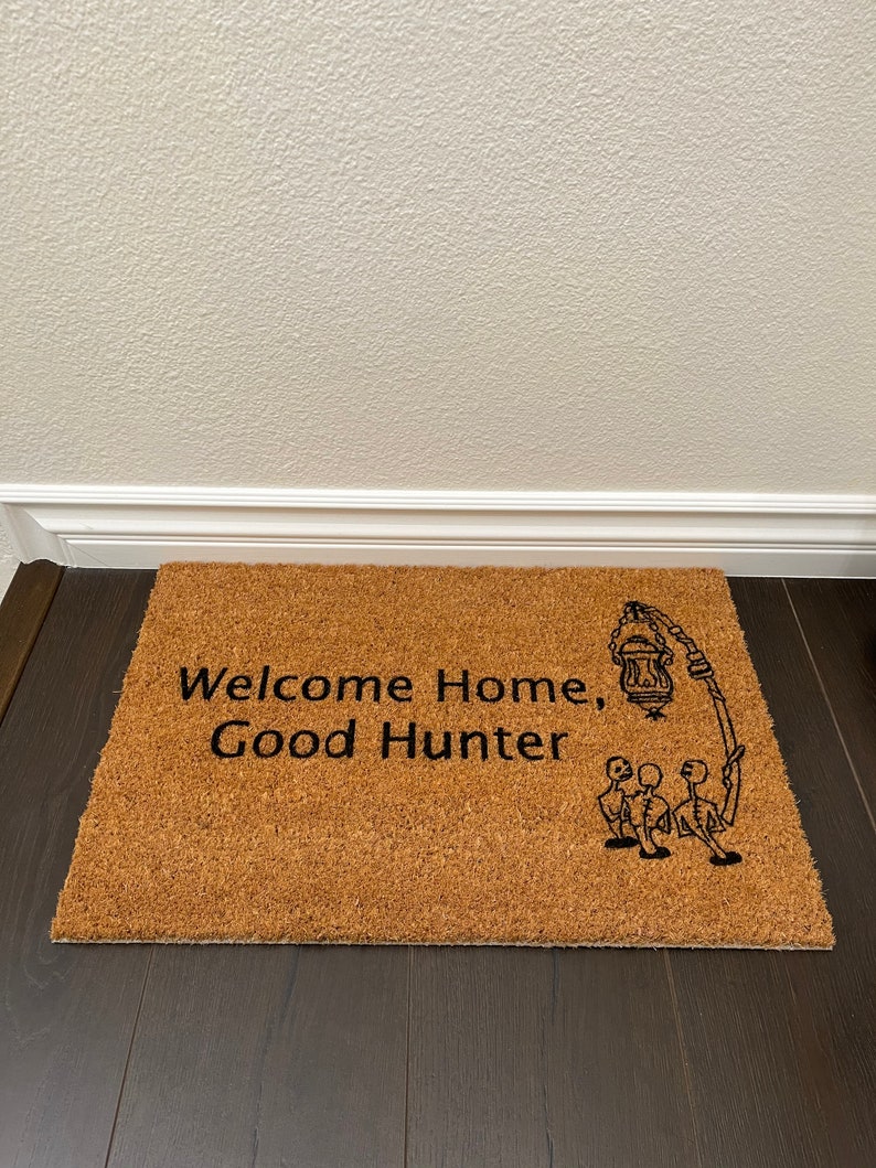 May include: A brown coir doormat with the text "Welcome Home, Good Hunter" and a black and white illustration of three figures holding a lantern.
