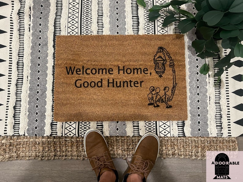 May include: A brown coir doormat with the text "Welcome Home, Good Hunter" and a black and white illustration of a lantern and three figures.