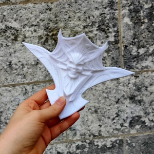 Op de afbeelding: Witte 3D-geprinte decoratieve wandhanger met een gedetailleerd ontwerp van een gezicht met hoorns en vleugels.