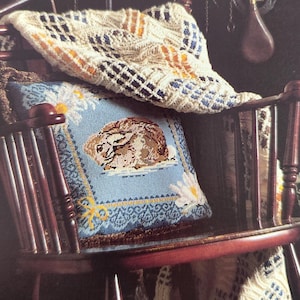 May include: A wooden rocking chair with a blue pillow featuring a rabbit design and a cream-colored blanket. The pillow has a light blue background with a rabbit illustration and daisy accents. The blanket has a geometric pattern with blue and orange accents.