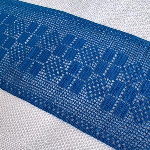 May include: A striking blue crocheted table runner with a geometric design. The runner is displayed on a white, textured surface. The intricate pattern and vibrant blue colour create a visually appealing contrast. The runner's dimensions are not visible.