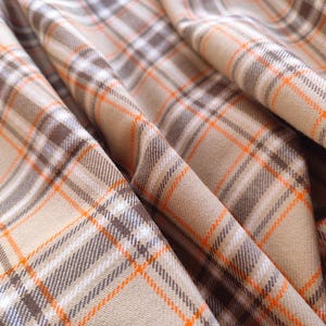 May include: Close-up of a folded fabric with a plaid pattern. The fabric features a beige base with intersecting lines in brown, white, and orange. The fabric is draped, showing the texture and folds of the material.