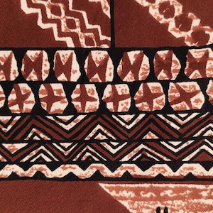 May include: Close-up of a textile with a repeating geometric pattern. The design features alternating rows of brown, black, and white shapes, including zigzags, crosses, and circles with cross-like details. The overall colour palette is warm, with shades of brown and cream.