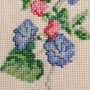 May include: Close-up of a cross-stitch floral design featuring blue and pink flowers with green leaves. The needlework is on a white fabric background, showcasing the detailed stitches and colour variations.