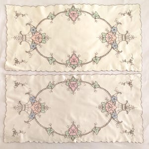 May include: Two off-white rectangular placemats with scalloped edges. Each features an embroidered floral design in pink, blue, and green, framed by a gray oval border. Delicate floral accents are scattered around the edges.