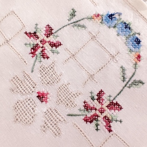 May include: A close-up of a white embroidered cloth with a floral design. The embroidery features red, pink, and blue flowers with green stems. The cloth has a grid-like pattern and a delicate, handmade appearance.