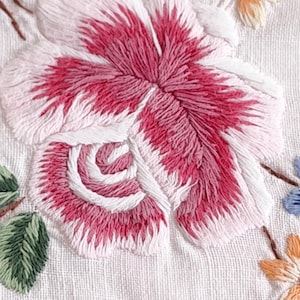 May include: Close-up of white fabric with floral embroidery. A large pink and white flower is the focal point, with detailed stitching. Green leaves and smaller blue and orange flowers complete the design.
