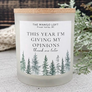 May include: A frosted glass candle jar with a light wood lid. The white label features the text "THE MANGO LOFT Fraser Valley, BC", "THIS YEAR I'M GIVING MY OPINIONS thank me later", and a watercolour illustration of green pine trees at the base.