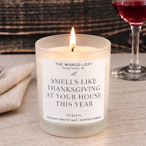 Peut inclure: Une bougie parfumée allumée dans un pot en verre transparent avec une étiquette blanche indiquant THE MANGO LOFT, FRASER VALLEY, BC, SMELLS LIKE THANKSGIVING AT YOUR HOUSE THIS YEAR, 226 g | 8 oz, COCONUT APRICOT WAX. Une mèche allumée.