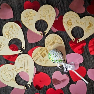 May include: Wooden heart-shaped decorations with engraved text and numbers, surrounded by red and pink heart confetti. The hearts feature phrases like "You + Me", "Six Seven", and "Friendship Level 67". A lollipop is in the center.