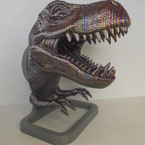 May include: A detailed, multi-colored Tyrannosaurus Rex bust with an open mouth, revealing sharp teeth. The dinosaur sculpture is mounted on a gray stand. The head and body have a textured, iridescent finish, creating a striking visual effect.