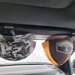 May include: Two baseball caps are clipped to a car visor. One cap has a digital camouflage pattern and the other is brown with a yellow mesh back. The brown cap has a white and black patch that reads "PDL Consulting Professional Engineering."