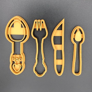 Puede incluir: Cuatro cortadores de galletas de metal de color dorado en forma de cuchara, tenedor, cuchillo y una cuchara pequeña. Los cortadores están sobre un fondo gris.