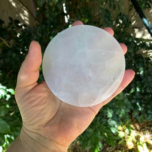 May include: A circular, translucent, white selenite charging plate. The plate is held in a hand, with a blurred green background of foliage. The selenite has a smooth surface and is used for cleansing crystals.