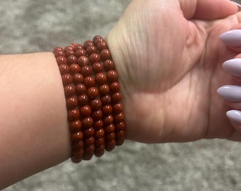 May include: A stack of six reddish-brown beaded bracelets. The beads are small and round, strung together on a thin, reddish string. The bracelets are worn on a wrist.