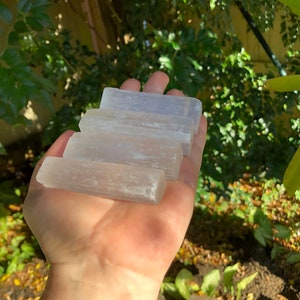May include: A collection of five selenite sticks, stacked on top of each other, in a hand. The sticks are white and translucent, with a slightly cloudy appearance.