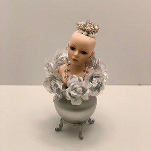 Puede incluir: Un busto de muñeca de porcelana con una corona plateada y un arreglo floral plateado y blanco. La muñeca lleva un collar plateado y el arreglo está en un jarrón plateado con tres patas.