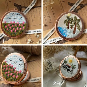May include: Four small, round, copper-coloured compact mirrors with embroidered designs. One features pink flowers, another palm trees, and a third a car on a road. The fourth mirror has a design of pink flowers and birds. Each mirror is labeled with a number.