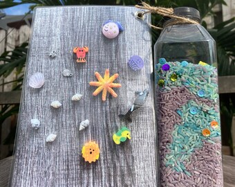 Puede incluir: Una botella de plástico transparente llena de arroz morado y azul y lentejuelas de colores. La botella está junto a una tabla de madera con varias decoraciones de temática marina, incluyendo una estrella de mar, un cangrejo, una concha y un pez.