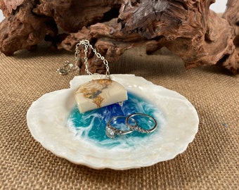 May include: A white seashell ring dish with blue and white resin waves. Two silver rings are resting in the dish. A gold and white necklace with a resin pendant is resting on the edge of the dish.