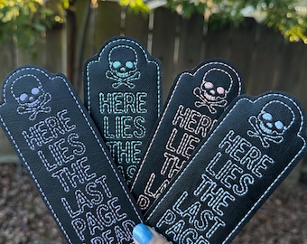 Puede incluir: Cuatro marcapáginas de cuero negro con un diseño de calavera y huesos cruzados y el texto "Here Lies The Last Page I Read". Los marcapáginas son de diferentes colores de hilo: rosa, azul, verde y blanco.