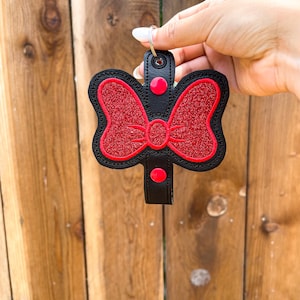 May include: A black leather keychain with a red glitter bow. The bow has two red buttons on the top and bottom.