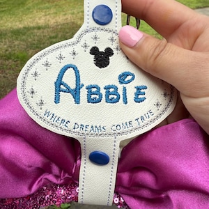May include: White leather keychain with a blue and silver embroidered design. The keychain features a Mickey Mouse silhouette, the name "Abbie", and the text "Where Dreams Come True".