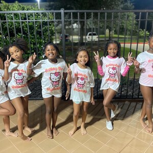 May include: Five girls wearing white t-shirts with a pink Hello Kitty graphic and the text "Birthday Girl" in orange. The girls are standing in front of a black metal fence.