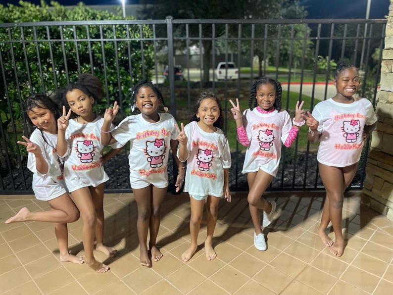 May include: Five girls wearing white t-shirts with a pink Hello Kitty graphic and the text "Birthday Girl" in orange. The girls are standing in front of a black metal fence.