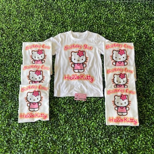 May include: Set of seven white t-shirts with Hello Kitty graphics. The shirts feature a pink Hello Kitty with a pink bow and the text "Birthday Girl" and "Birthday Crew" in orange.