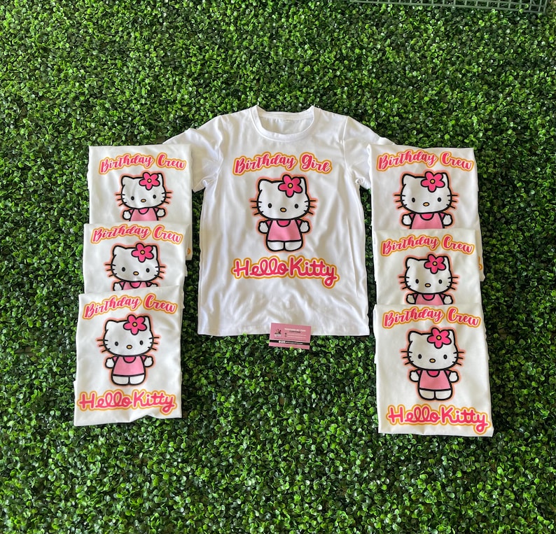 May include: Set of seven white t-shirts with Hello Kitty graphics. The shirts feature a pink Hello Kitty with a pink bow and the text "Birthday Girl" and "Birthday Crew" in orange.