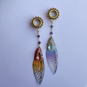 May include: A pair of gold-toned ear gauges with iridescent wings and crystal accents. The wings are a gradient of blue, purple, and pink. The crystals are clear and faceted.