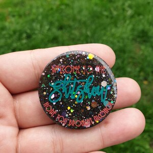 May include: A round black resin pin with glitter embedded in the resin. The pin says "Welcome to the shitshow. Hope you brought alcohol." in pink and teal lettering.
